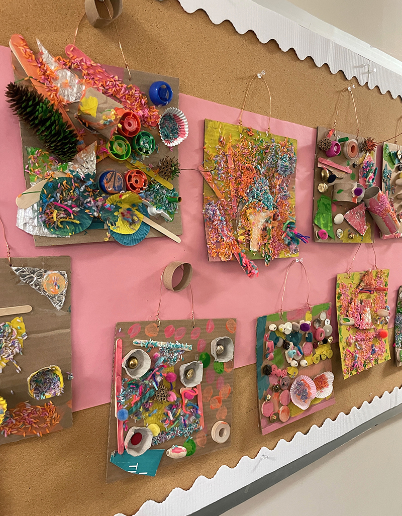 Recycled collages made by preschoolers hanging up on a wall in a hallway for display.