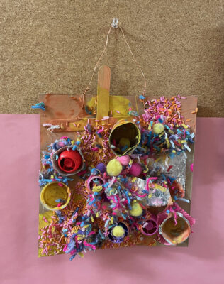 A finished recycled collage made by a preschooler using cardboard, tp rolls, glue, paint, and yarn