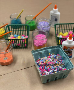Setting up for part 2 of recycled collages with preschoolers, using yarn, dyed rice, tempera paint, and pom-poms.