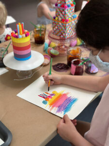 Childr at a long table lined with an array of recycled cakes using oil pastels and liquid watercolor.