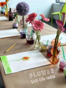 Table full with small bud vases, flowers, paper, pencils, and watercolors.