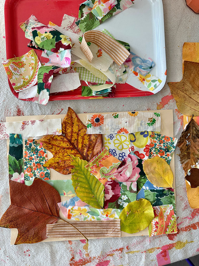 Finished mixed-media collage using leaves and fabric on a table next to a tray of fabric scraps.