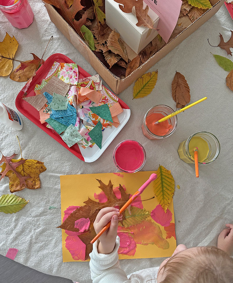 A young child is painting with temperas and collage with leaves and fabric on colored construction paper.