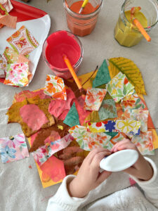 A young child is glueing leaves and fabric strips to her painted collage.