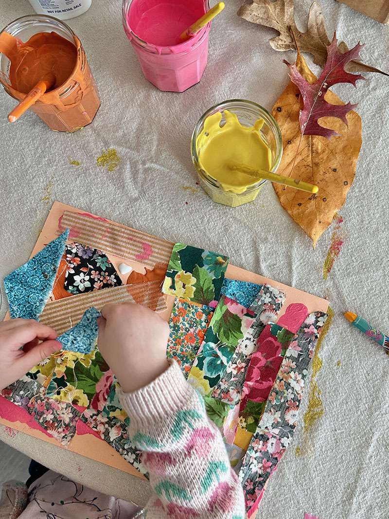 A young child is creating a collage with leaves and fabric on colored construction paper.