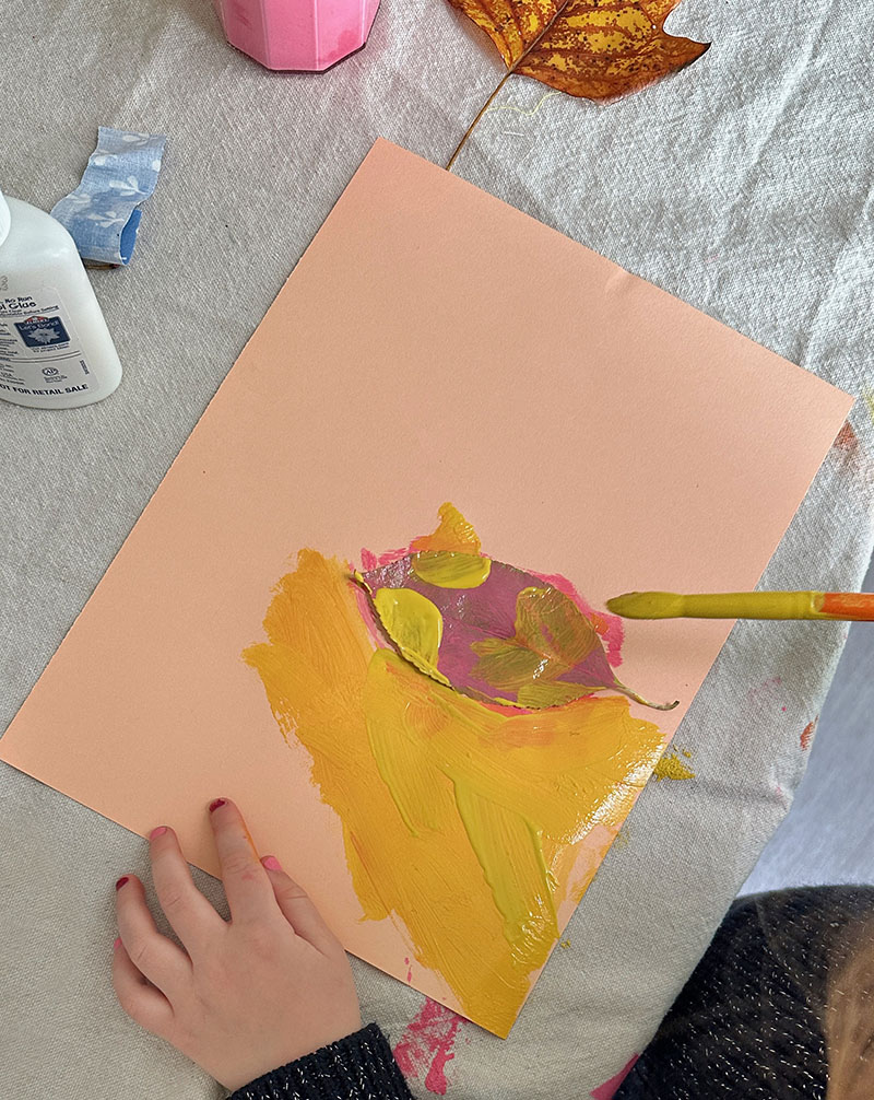 Child painting with tempera paints on top of construction paper and leaves.