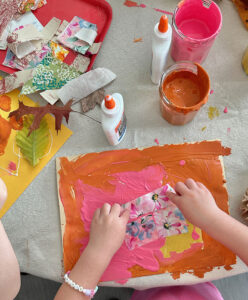 Child adding fabric scraps to her painting, with leaves and glue on the table.