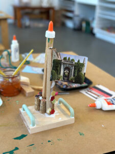 A wood structure made by a 7-yr old using corks, buttons, tops, and magazine clippings.