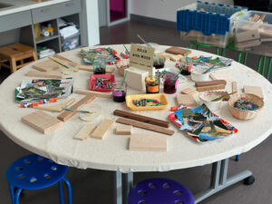 A table set up for children to make art with wood pieces, magazine clippings, collage materials, glue, and liquid watercolor.