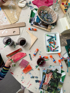 Child painting wood pieces with liquid watercolor on a table set up with other collage material and oil pastels.