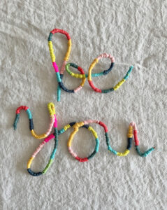 The words "Be You" are made with pipe cleaners and yarn and are colorfully displayed on a table.