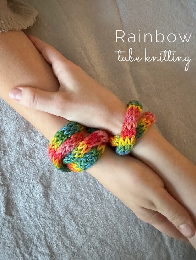 Rainbow tube knitted bracelents wrapped around two hands holding each other.