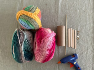 Yarn, tp roll, popsicle sticks, buttons, and a glue gun are are on a table ready to made into a tube knitting loom.