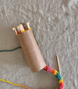 Rainbow colored tube knitting on a table with a tp roll loom.