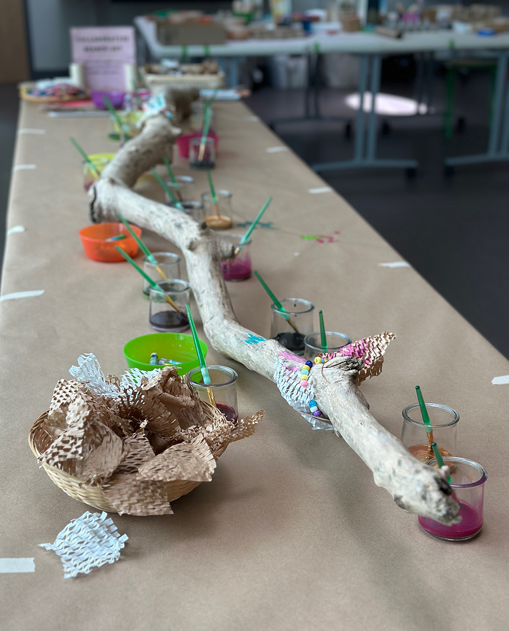 A big driftwood branch lays on a table with jars of paint surrounding it, ready for children to come and embellish.