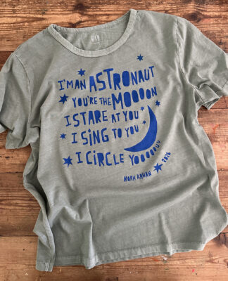 A newly printed t-shirt lies on a table showing off the freezer paper stencil technique using bright blue ink on a muted green shirt with a Noah Kahan quote from his song Orbiter.