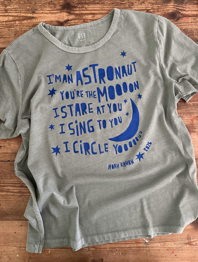 A newly printed t-shirt lies on a table showing off the freezer paper stencil technique using bright blue ink on a muted green shirt with a Noah Kahan quote from his song Orbiter.